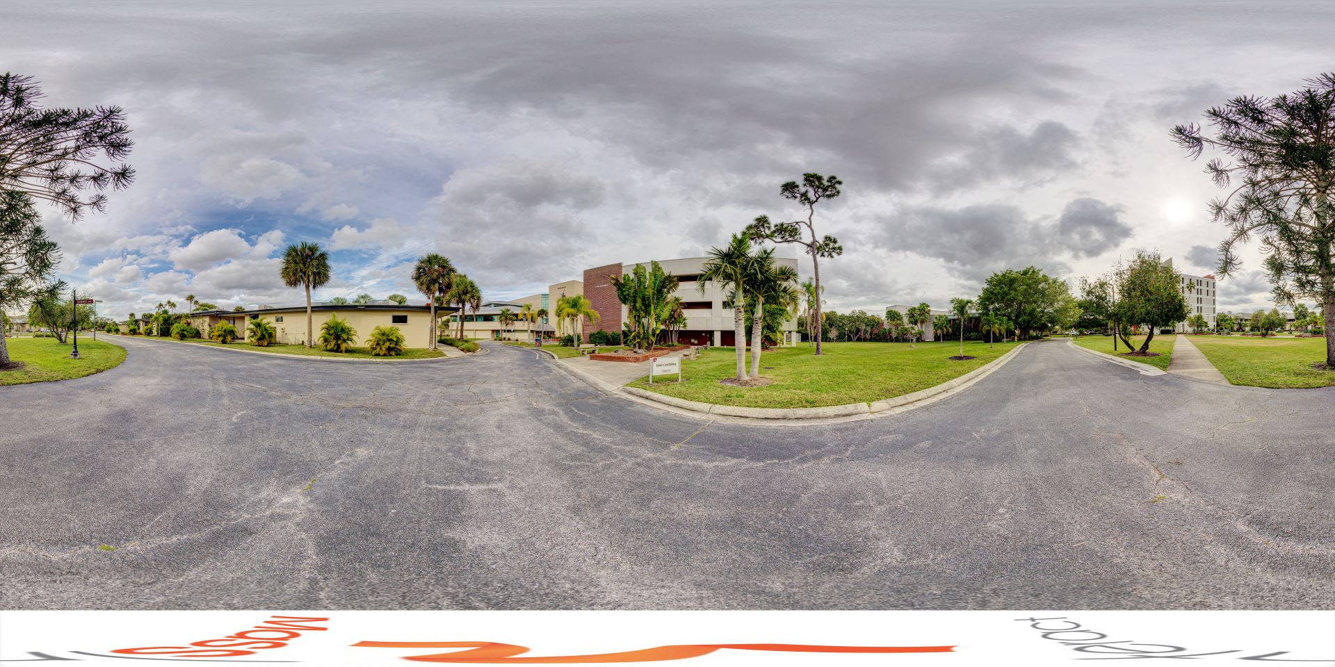 Panoramic view of a university campus with the Link Building in the center, surrounded by lush green lawns and palm trees, under a dramatic cloudy sky